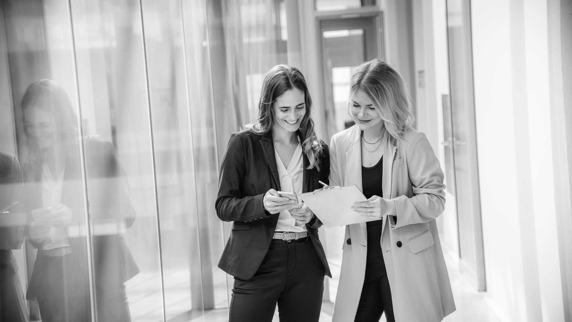 Sarah Hood und Carmen Holtkamp bei der Arbeit. Beide schauen auf ein Dokument und lächeln dabei. Das Foto ist schwarz / weiß