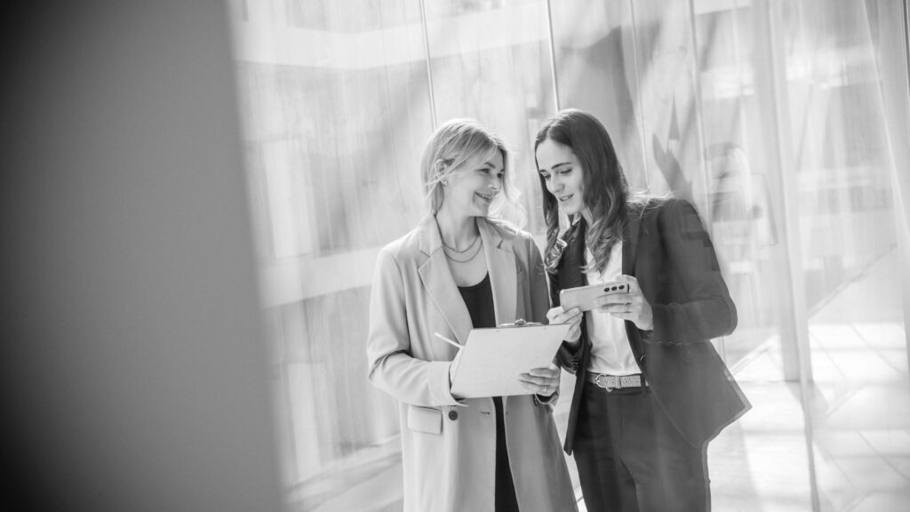 Sarah Hood und Carmen Holtkamp bei der Arbeit bei traditionvonmorgen. Sie haben ein Handy und ein Klemmbrett dabei und lächeln sich an. Das Foto ist schwarz / weiß